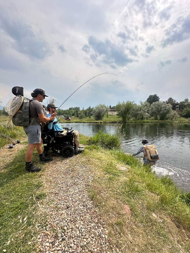 Support team member and participant in a power chair fighting a fish from the riverbank.