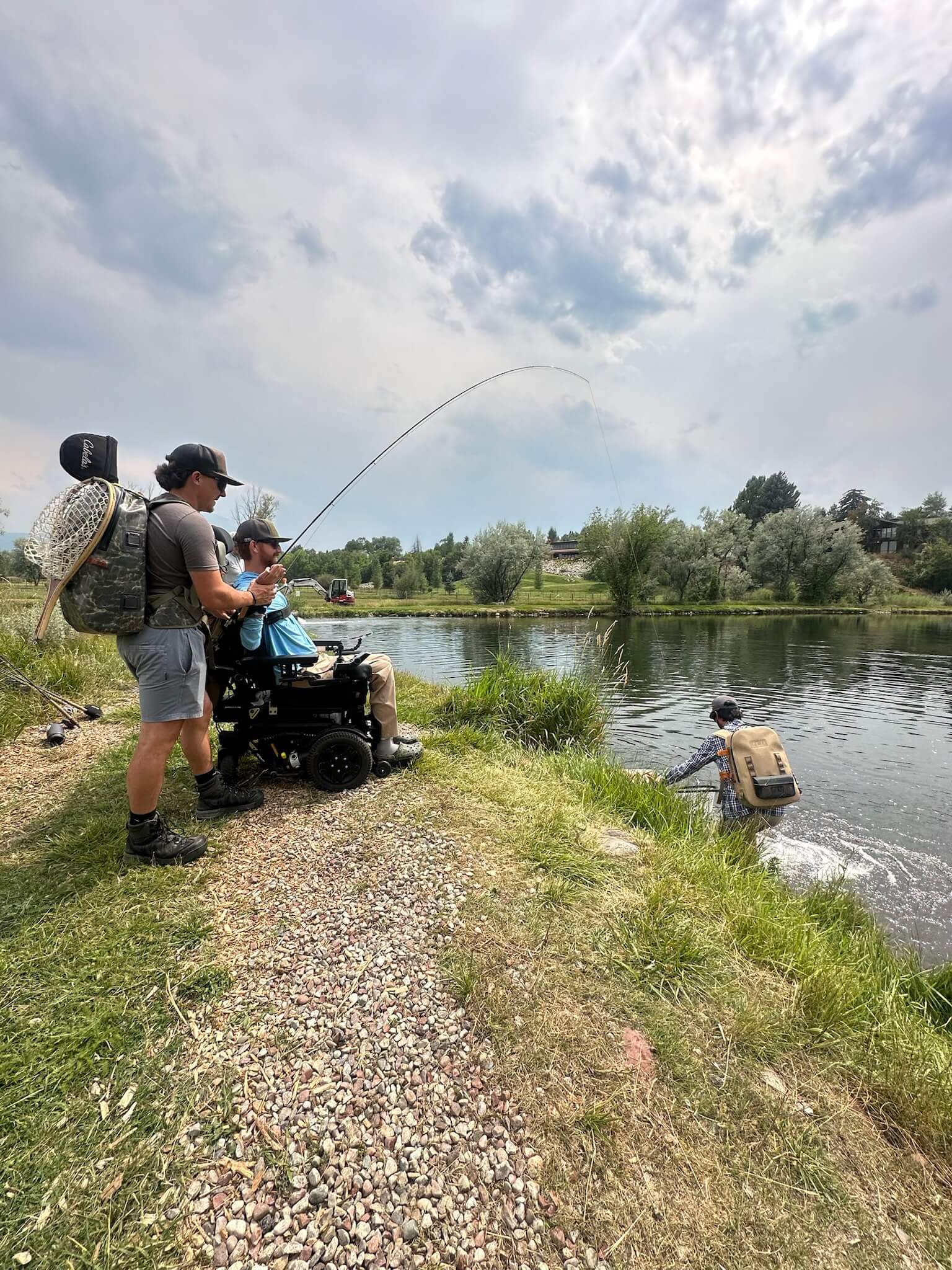 Support team member and participant in a power chair fighting a fish from the riverbank.