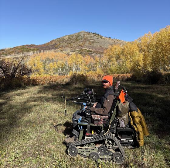 Hunter in blaze orange seated in a tracked wheelchair in a meadow below golden aspens.