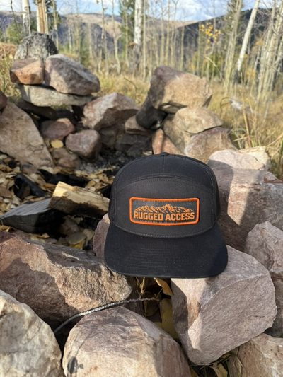 Black 7-panel hat with an orange-bordered Rugged Access mountain patch resting on a ring of fire-pit rocks.