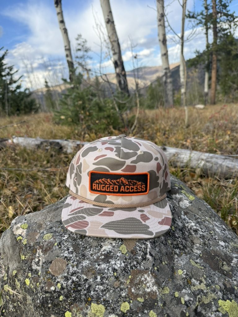 Desert duck camo rope hat with an orange-and-black Rugged Access mountain patch, sitting on a lichen-covered boulder.