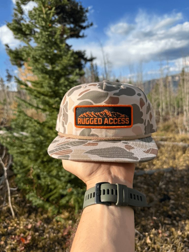 Hand holding up the camo rope hat in front of a pine tree, patch facing the camera.
